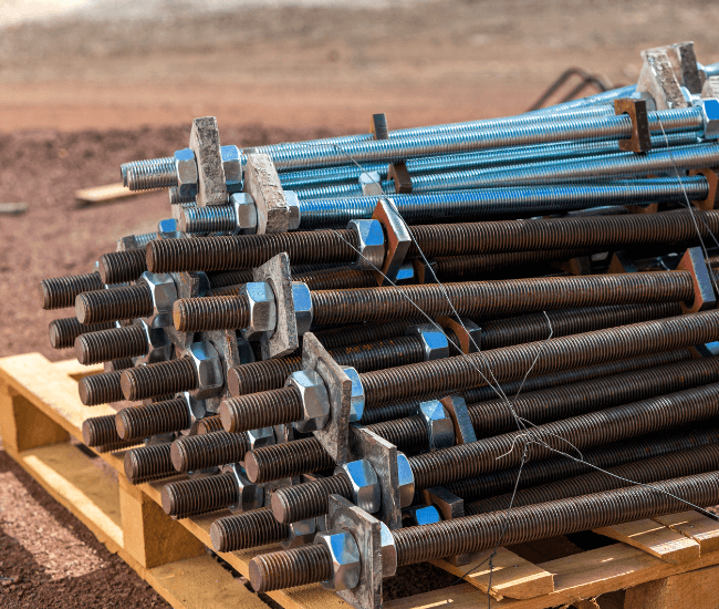 Stack of threaded rebar with hex nuts, some with protective blue caps, arranged on a wooden pallet at a construction site.