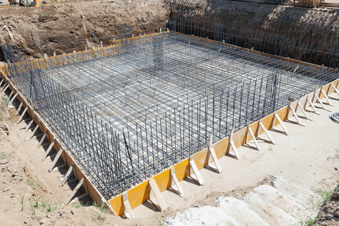Reinforcement steel bars forming a grid pattern for a foundation, with wooden formwork and trench shields around the excavation site, under bright daylight.