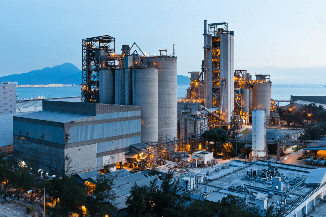 Twilight view of an industrial cement plant with towering silos and structures illuminated by lights, with a backdrop of mountains and a clear sky.