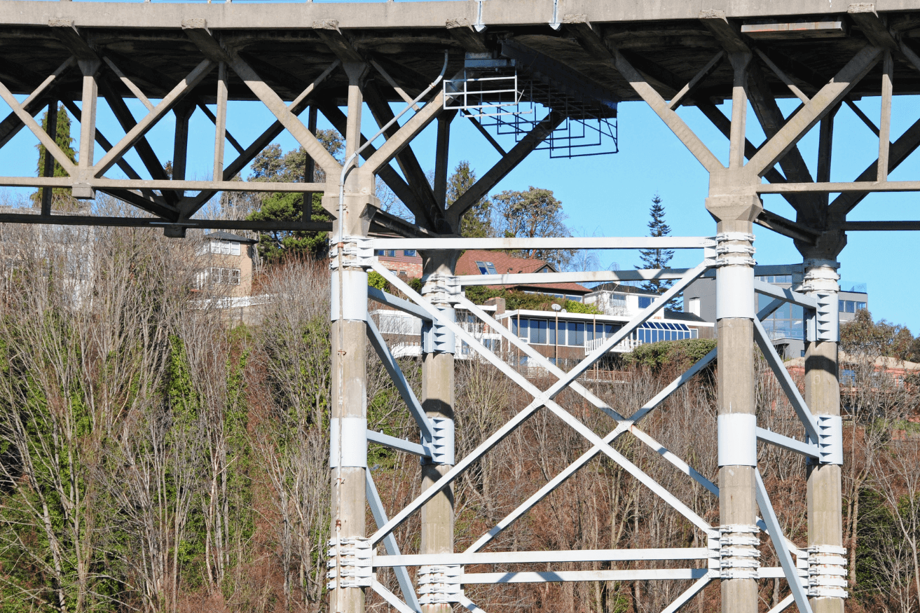 Underneath view of a complex steel bridge structure with diagonal braces, showing detailed engineering against a backdrop of trees and a house on a hill.