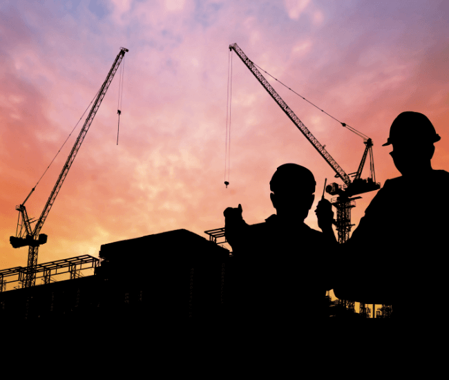 Silhouettes of two construction workers wearing hard hats with tower cranes in the background, against a vivid sunset sky.