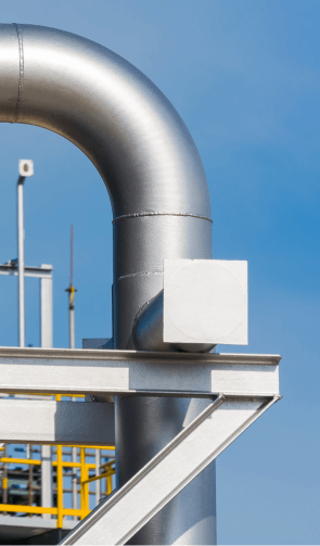 Close-up of a metallic elbow pipe with a blank square identification plate against a clear blue sky, with industrial structures in the background.