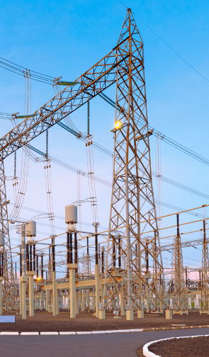 The image depicts a high-voltage electrical substation with several transmission towers and power lines. The substation includes multiple insulators, transformers, and other electrical components crucial for distributing electricity. The towers and lines are arranged in a complex network, illustrating the infrastructure required for power transmission. The sky is clear, suggesting the photo was taken during daylight hours, and a paved road or path is visible in the foreground.