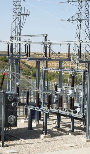 The image shows various structures and equipment within an electrical substation. Visible are multiple high-voltage insulators, circuit breakers, and busbars, interconnected by thick cables and supported by metal frameworks. Transmission towers and power lines are also present in the background, highlighting the substation's role in distributing electricity. The area is well-organized with gravel ground covering and fencing, ensuring safety and proper maintenance access. The surrounding landscape includes grassy areas and a clear sky, indicating daytime with good weather conditions.