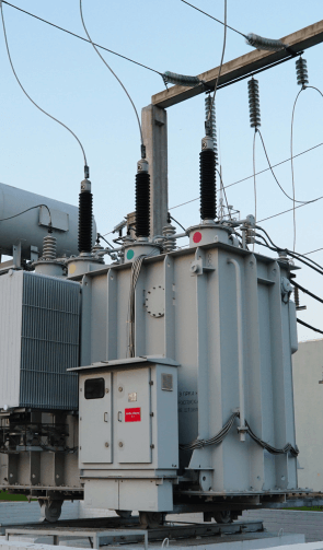 The image shows a large electrical transformer at a substation. The transformer is equipped with multiple high-voltage insulators, bushings, and cooling fins. It is connected to overhead power lines and surrounded by a network of cables and equipment essential for electricity distribution. The transformer is mounted on a concrete base, and various control panels and safety signs are visible on its exterior. The clear sky suggests it is daytime, providing good visibility of the detailed components of the transformer and the infrastructure around it.