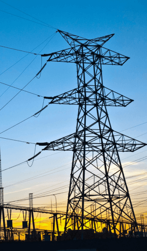 The image depicts a high-voltage transmission tower silhouetted against the sky during sunset or sunrise. The tower supports multiple power lines and is part of a larger network of electrical infrastructure visible in the background. The intricate lattice structure of the tower is highlighted by the contrasting light, creating a striking visual effect. The sky features a gradient of colors, transitioning from deep blue to warm yellow and orange hues near the horizon, indicating the time of day.