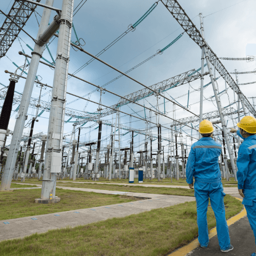 Two engineers in safety helmets and blue uniforms inspect a high-voltage electrical substation with steel towers and overhead power lines on a cloudy day.